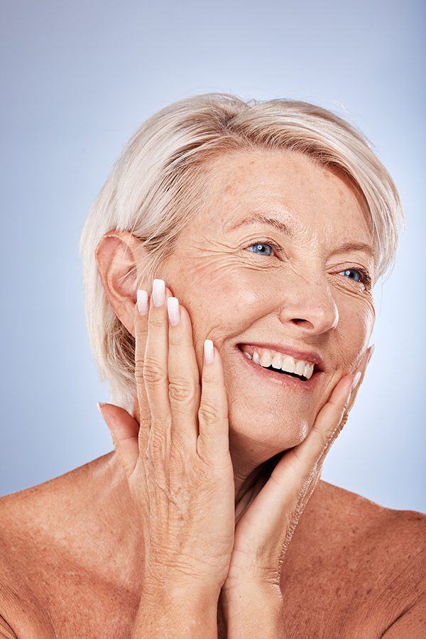 A smiling older woman with short, white hair gently touching her face with both hands, showcasing her radiant skin against a light blue background.