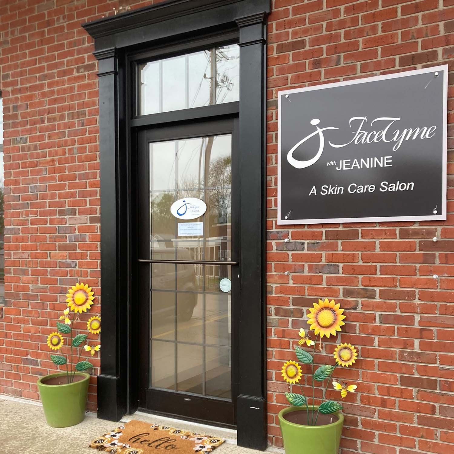 An entrance to FaceTyme with Jeanine, A Skin Care Salon, featuring a brick wall, a black-framed glass door, and a sign with the salon's name.