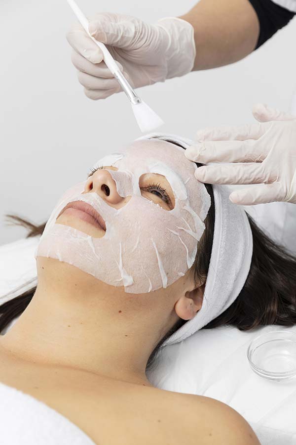 A woman receiving a facial treatment with a sheet mask applied, while a professional applies additional product with a brush.