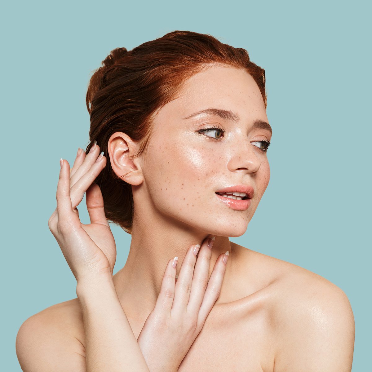 A young woman with fair skin and red hair poses gracefully with her hands touching her face and neck against a light blue background.